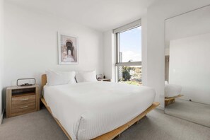 A bedroom with a large bed, a bedside table, a nightstand, a window with a view, and a framed picture on the wall. - Skyline-View 2-Bed Apartment Near Flemington (Flemington)
