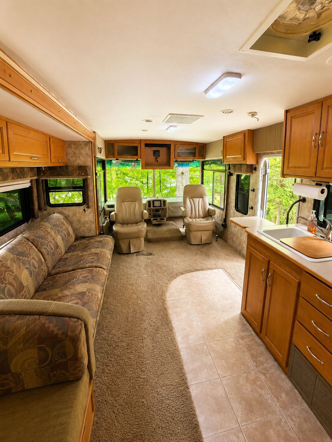 Compact camperinrichting met een bank, twee stoelen, een kleine keukenhoek en een raam met uitzicht op groen.