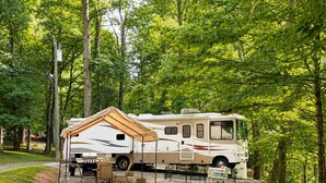 Un veicolo ricreativo (camper) parcheggiato in un’area sterrata, circondato da alberi, con un tavolo e sedie da picnic coperti nelle vicinanze.