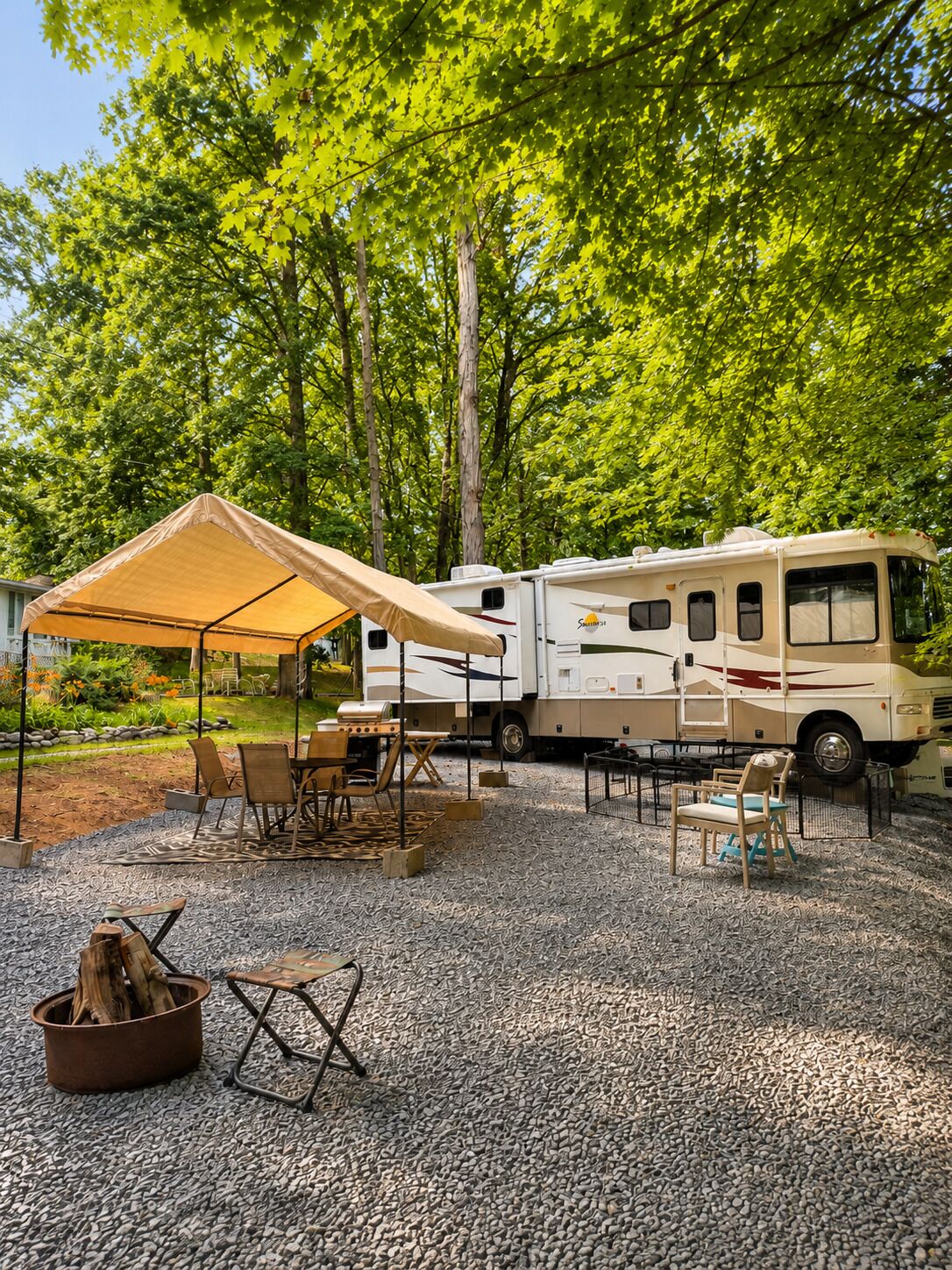 An RV parked in a gravel area with a canopy, surrounded by trees and outdoor furniture.
