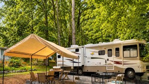 Un camper parcheggiato su un'area sterrata con un tendale, circondato da alberi e mobili da esterno.