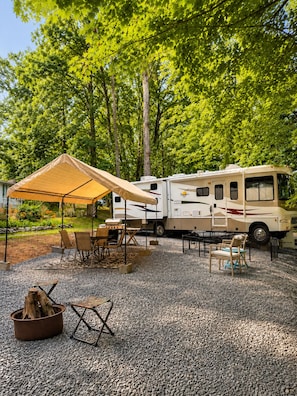 Un camping-car est stationné sur une aire de gravier, sous un auvent, et est entouré d’arbres et de meubles de jardin.