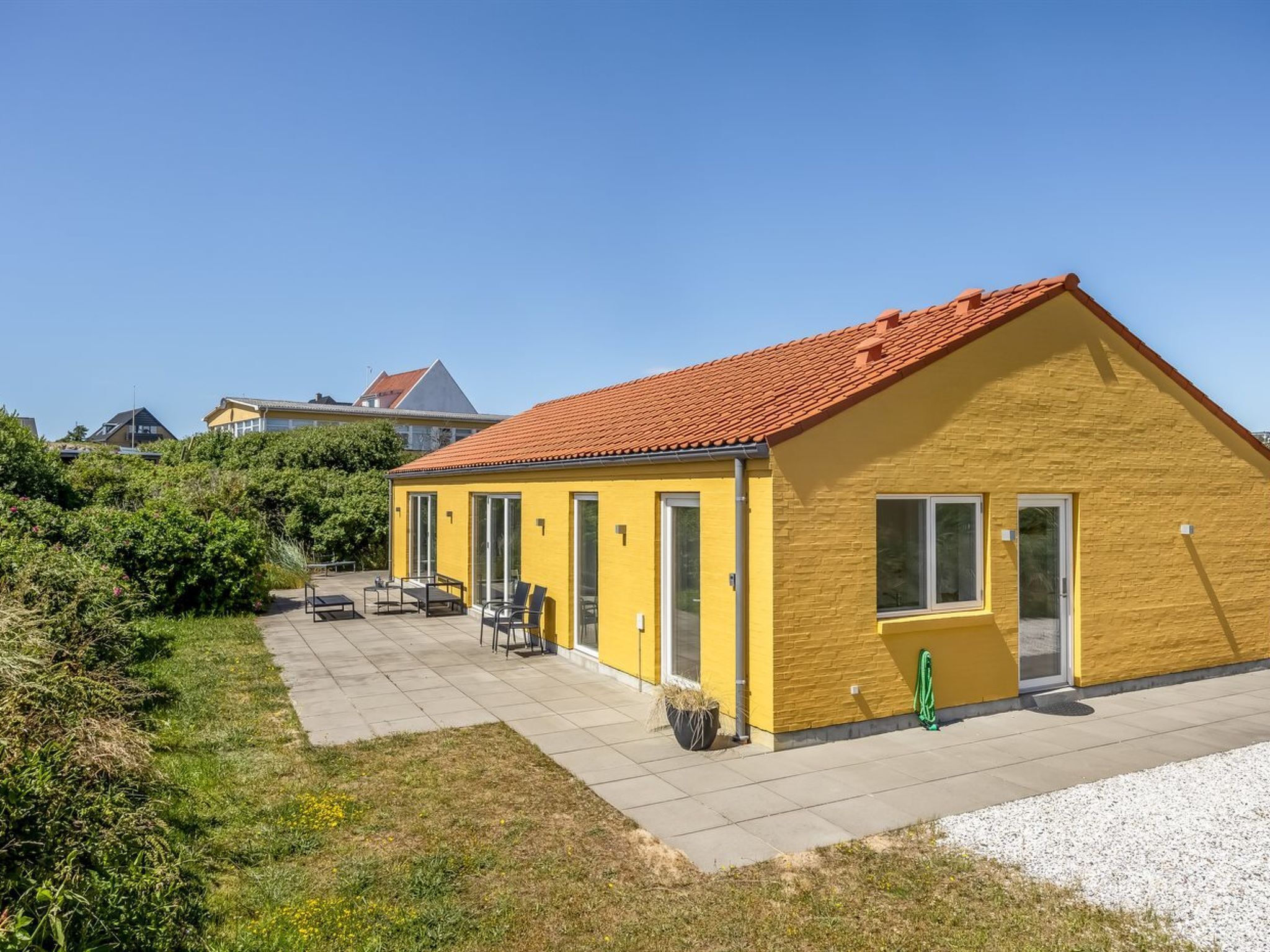Un bâtiment jaune avec un toit recouvert de tuiles rouges, une terrasse avec des meubles d’extérieur et un espace recouvert de gravier.