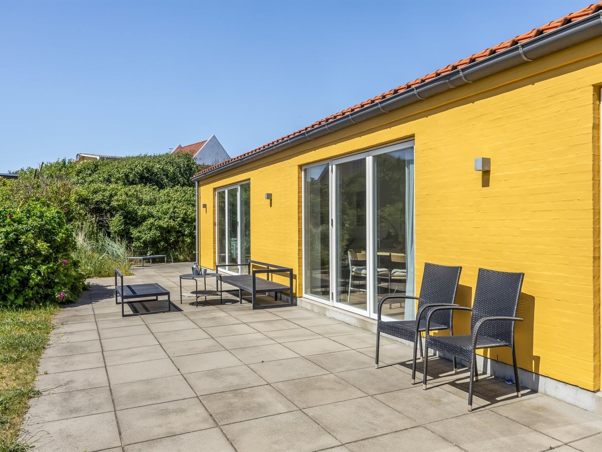 Un bâtiment jaune doté de grandes portes coulissantes en verre, une terrasse avec des meubles d’extérieur et un ciel dégagé.