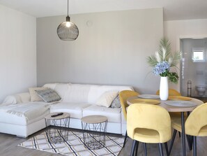Ein modernes Wohnzimmer mit einer Couch, zwei Beistelltischen, einem Esstisch mit Stühlen, einer Pendelleuchte und einer Topfpflanze.