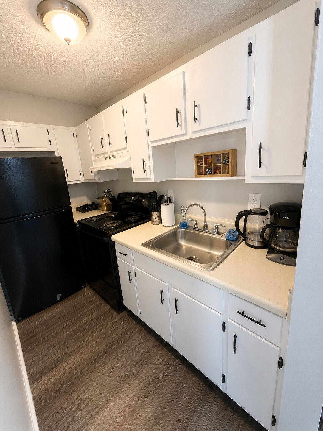 A compact kitchen with white cabinets, a black refrigerator, a stove, a sink, and a coffee maker.