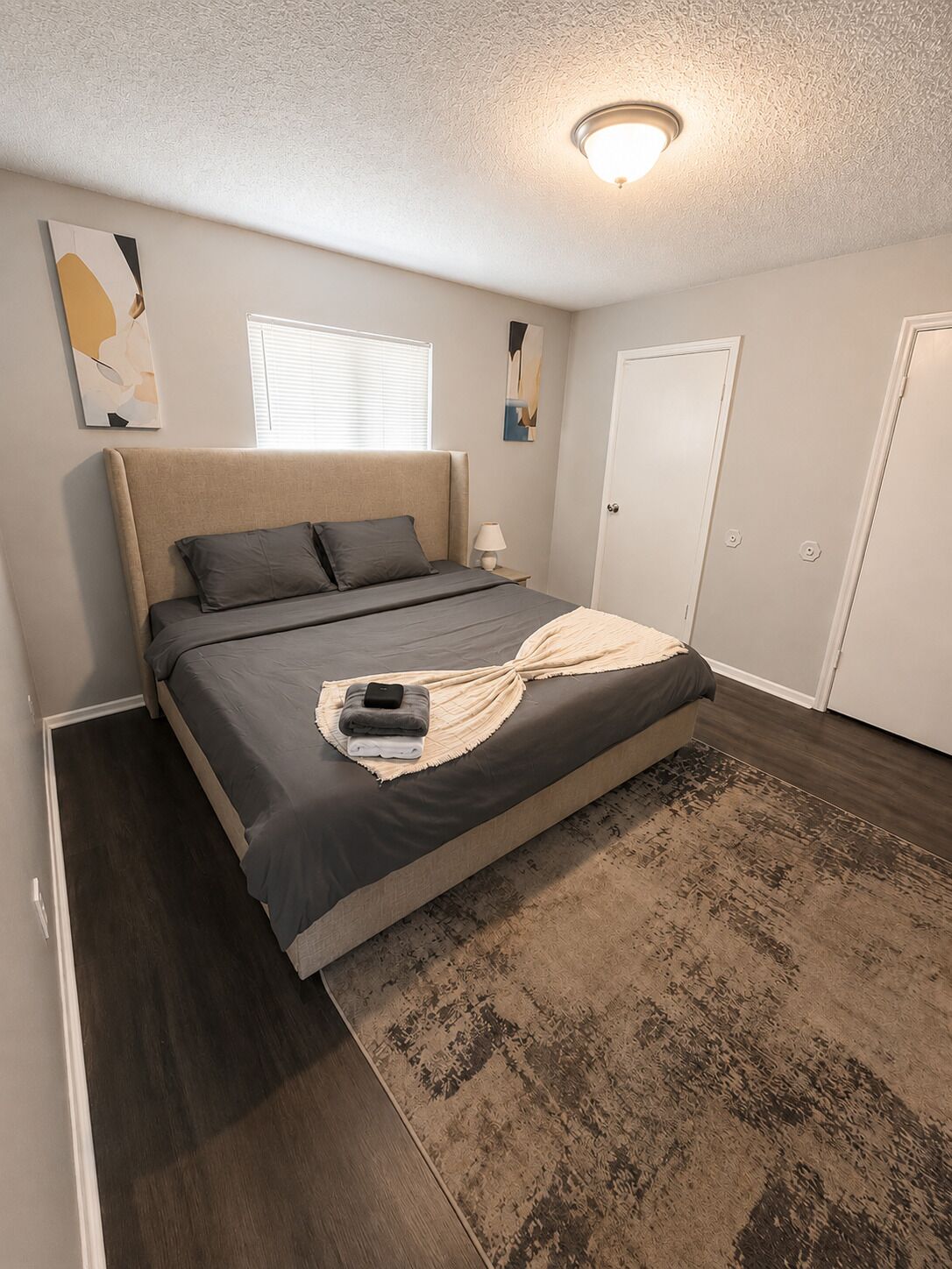 A bedroom with a bed, a bedside table, a lamp, a window, and a door.