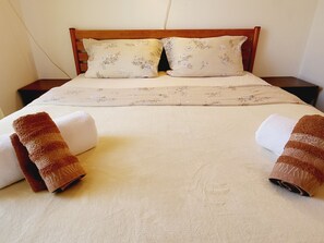 Een opgemaakt bed met bloemenkussens en een passend dekbedovertrek, twee handdoeken aan weerszijden, en een houten hoofdbord.