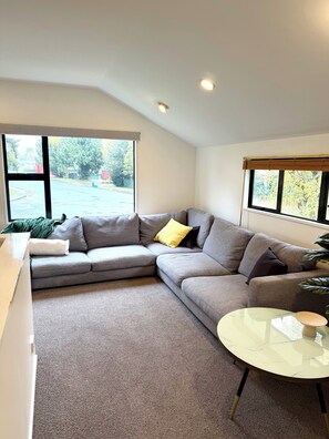 A modern living room with a grey sectional sofa, a round coffee table, and large windows offering a view of the outdoors. - Snowfed House - perfect for 2 families or large groups | sleeps 9 (Methven)