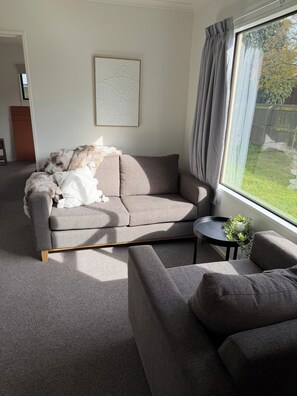 A living room with a grey sofa, a smaller grey armchair, a coffee table with a plant, and a window with curtains. - Snowfed House - perfect for 2 families or large groups | sleeps 9 (Methven)
