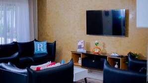 Een woonkamer met een flatscreen televisie aan de muur, een bank met een decoratief kussen, een salontafel, en een bijzettafeltje met een plant.