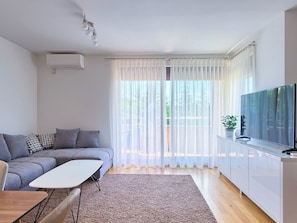 A modern living room with a grey sofa, a white coffee table, a flat-screen TV, and a large window with sheer curtains. - Mara spacious apartment with pool by Interhome (Medulin/Ližnjan)