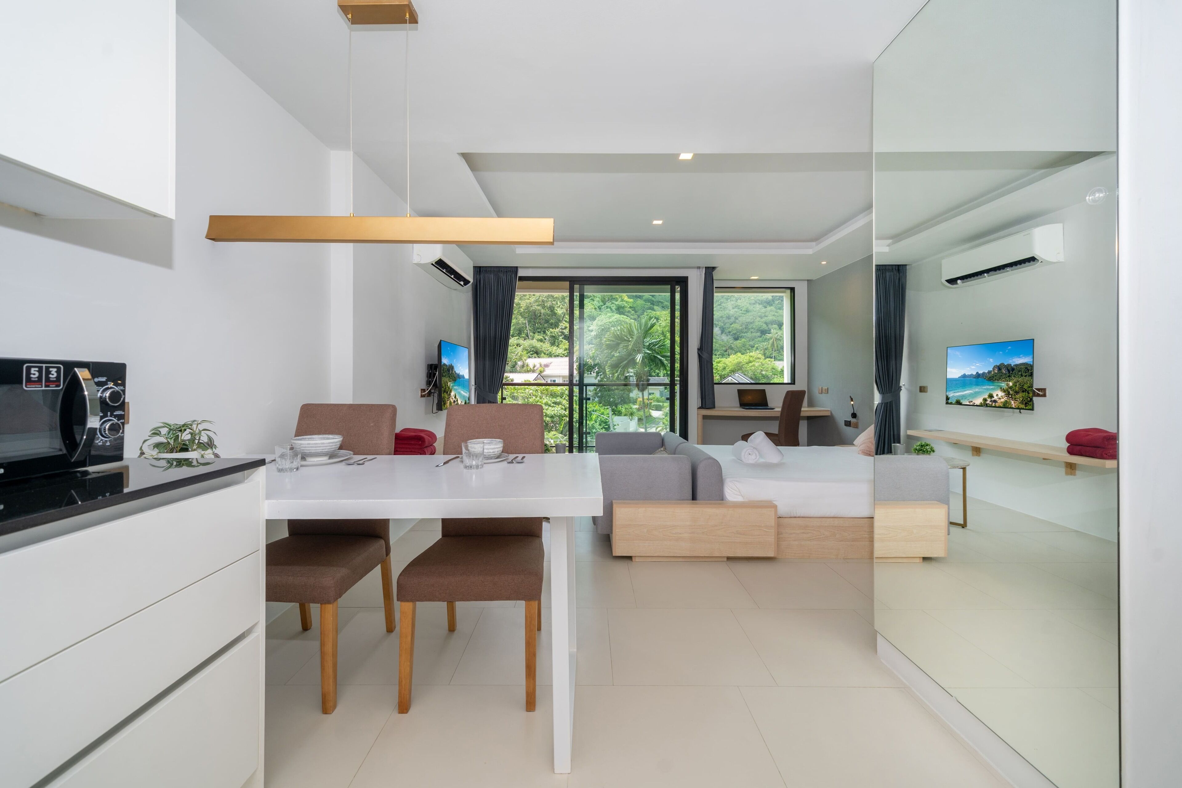 A modern living space with a dining area, a sofa, a TV, and a view of greenery.