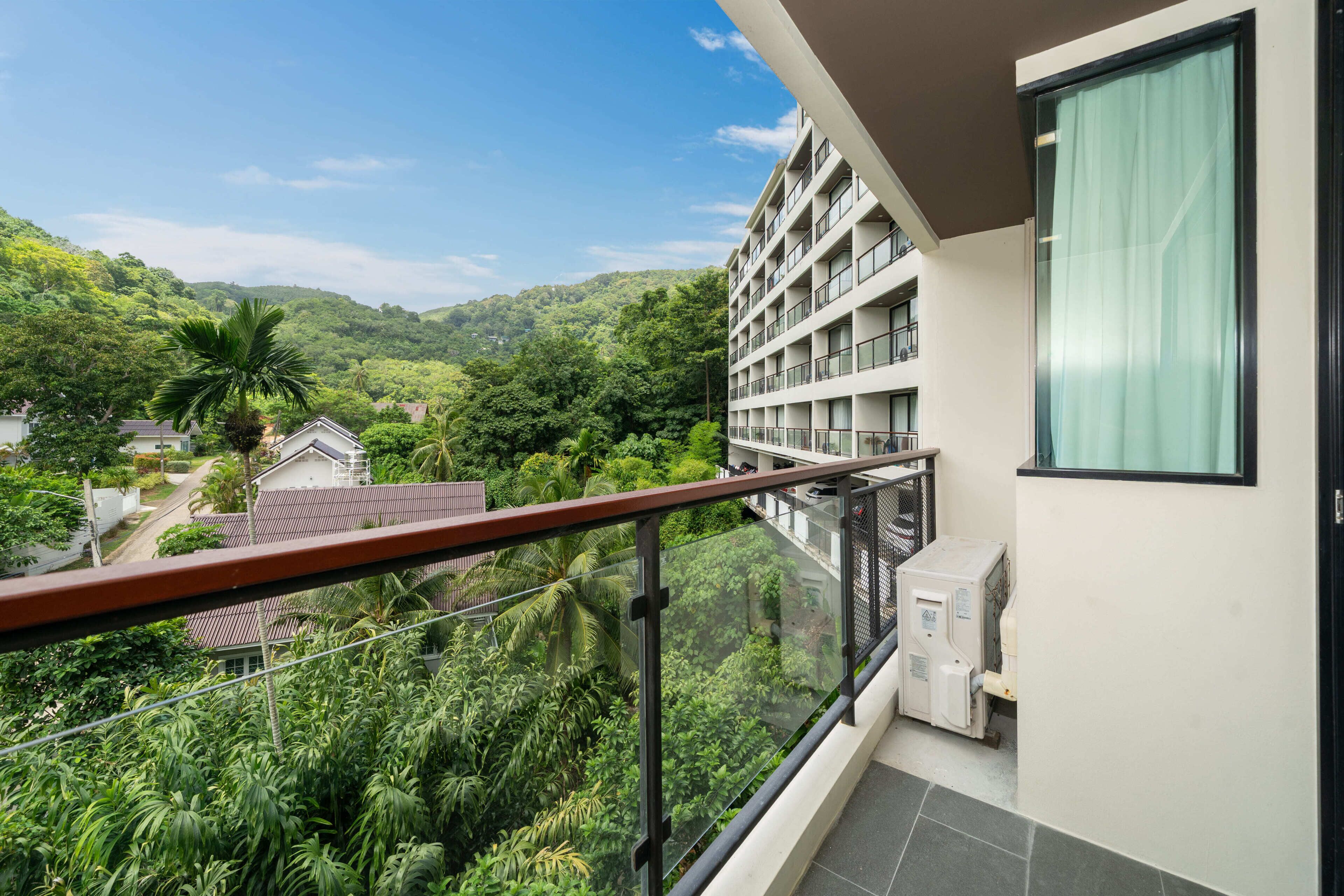 A modern hotel balcony with a glass railing overlooking a lush green landscape and a hillside.