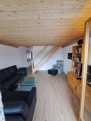 A living room with a wooden ceiling, a black leather sofa, a glass coffee table, a plaid armchair, and a wooden bookshelf. - I dr Rehwiesa B31 Appartement/Fewo, Bad, WC, 4-Bettwohnung by Interhome (Arosa)