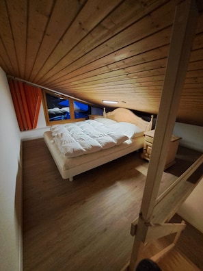 A bedroom with a wooden sloped ceiling, a bed with white bedding, a nightstand, and a window with curtains. - I dr Rehwiesa B31 Appartement/Fewo, Bad, WC, 4-Bettwohnung by Interhome (Arosa)