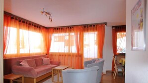 Ein Wohnzimmer mit einer Couch, einem kleinen Tisch und einem Sessel. Große Fenster mit orangenen Vorhängen.