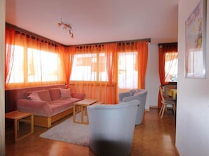 Ein Wohnzimmer mit einer Couch, einem kleinen Tisch und einem Sessel. Große Fenster mit orangenen Vorhängen.