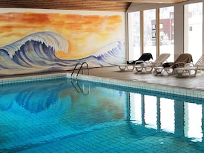 Ein Hallenbad mit einer Wellenmalerei an der Wand, Liegestühle und freier Sicht nach draußen.