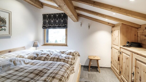 A bedroom with a bed, a window with a patterned valance, a small stool, and wooden cabinets. - Chalet Steinbock by Arosa Holiday Appartement/Fewo, Bad, WC, 3 Schlafräume by Interhome (Arosa)