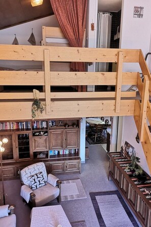 Een loft-achtige kamer met een houten trap, een zithoek met een gestreept fauteuil, een boekenkast en een eettafel.