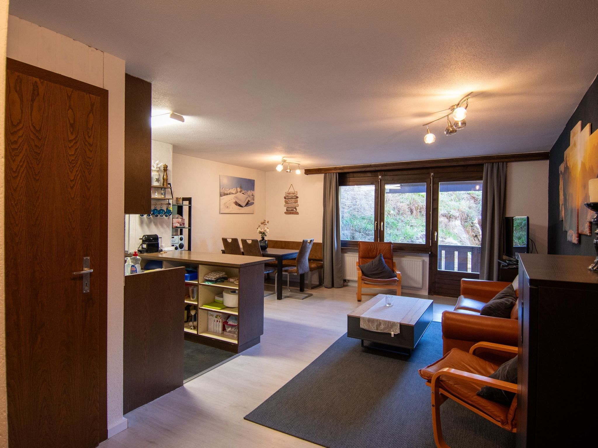 Ein gemütliches Wohnzimmer mit einer Couch, einem Couchtisch und einem Essbereich mit Stühlen und Tisch. Es gibt eine Küchenzeile mit Schränken und einem Kühlschrank. Der Raum hat große Fenster mit Vorhängen und Blick ins Freie.