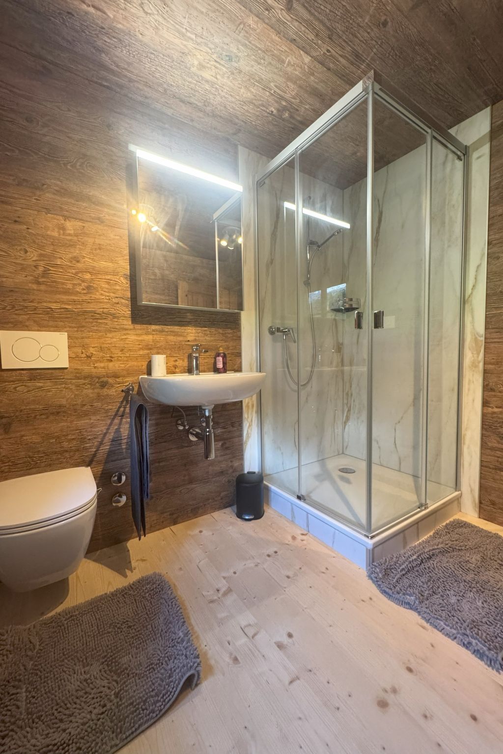 Ein Badezimmer mit Holzwand, einer Duschkabine aus Glas, einem weißen Waschbecken und einer Toilette.
