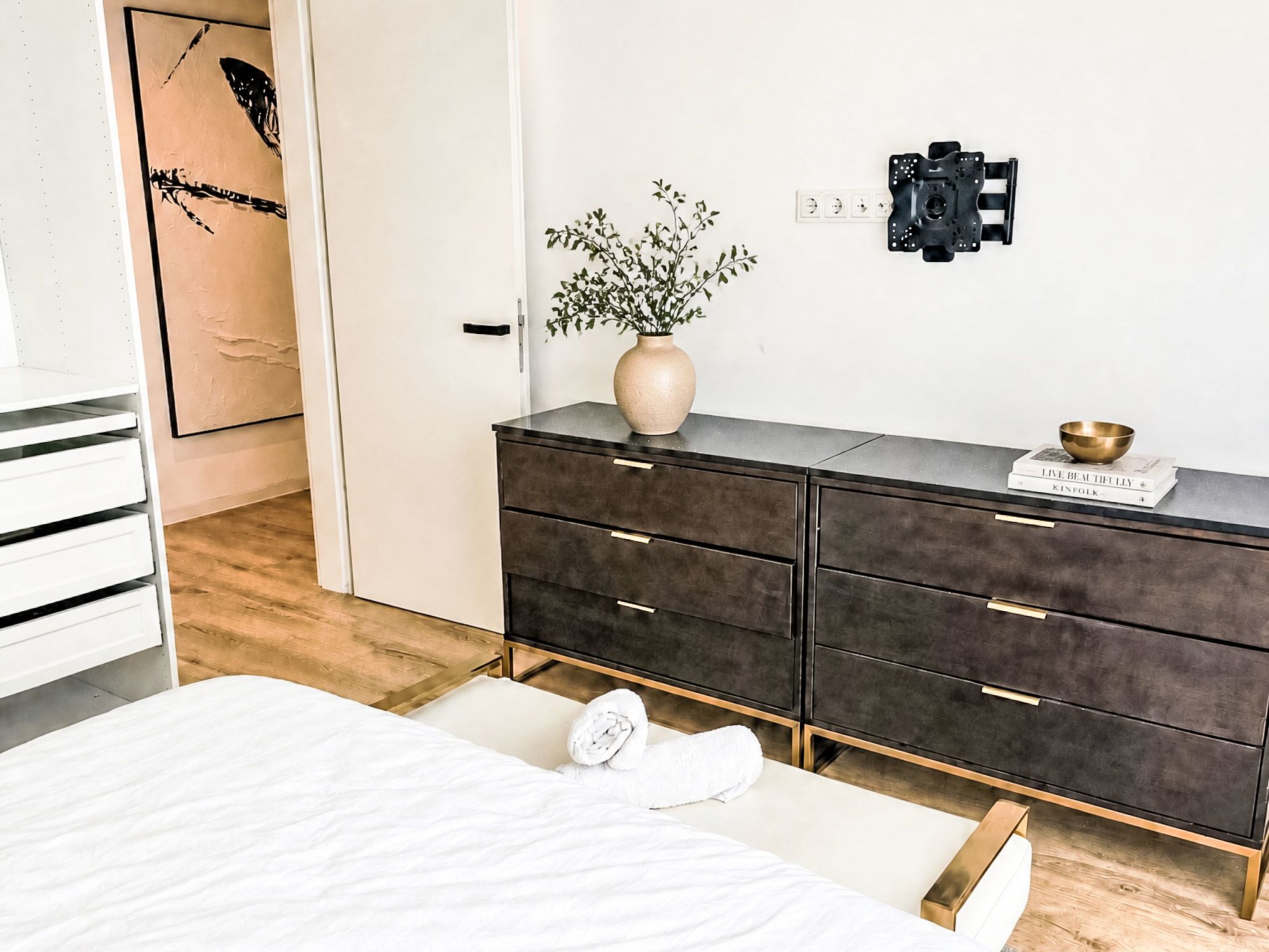 Ein Schlafzimmer mit einem Bett, einer Kommode mit Schubladen, einer Vase mit Pflanzen und einer Wandleuchte.