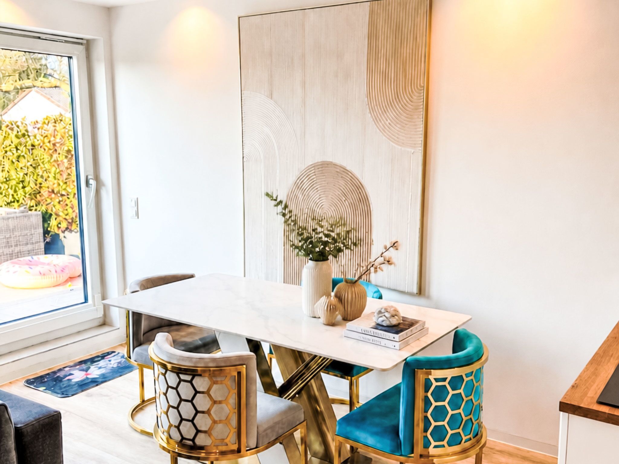A dining area with a white table, two chairs with gold and blue upholstery, a vase with flowers, and a large abstract painting on the wall.
