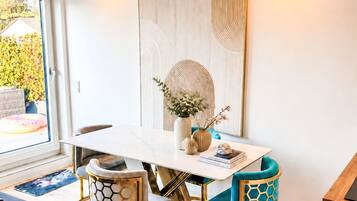 A dining area with a white table, two chairs with gold and blue upholstery, a vase with flowers, and a large abstract painting on the wall.