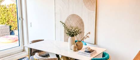 A dining area with a white table, two chairs with gold and blue upholstery, a vase with flowers, and a large abstract painting on the wall.