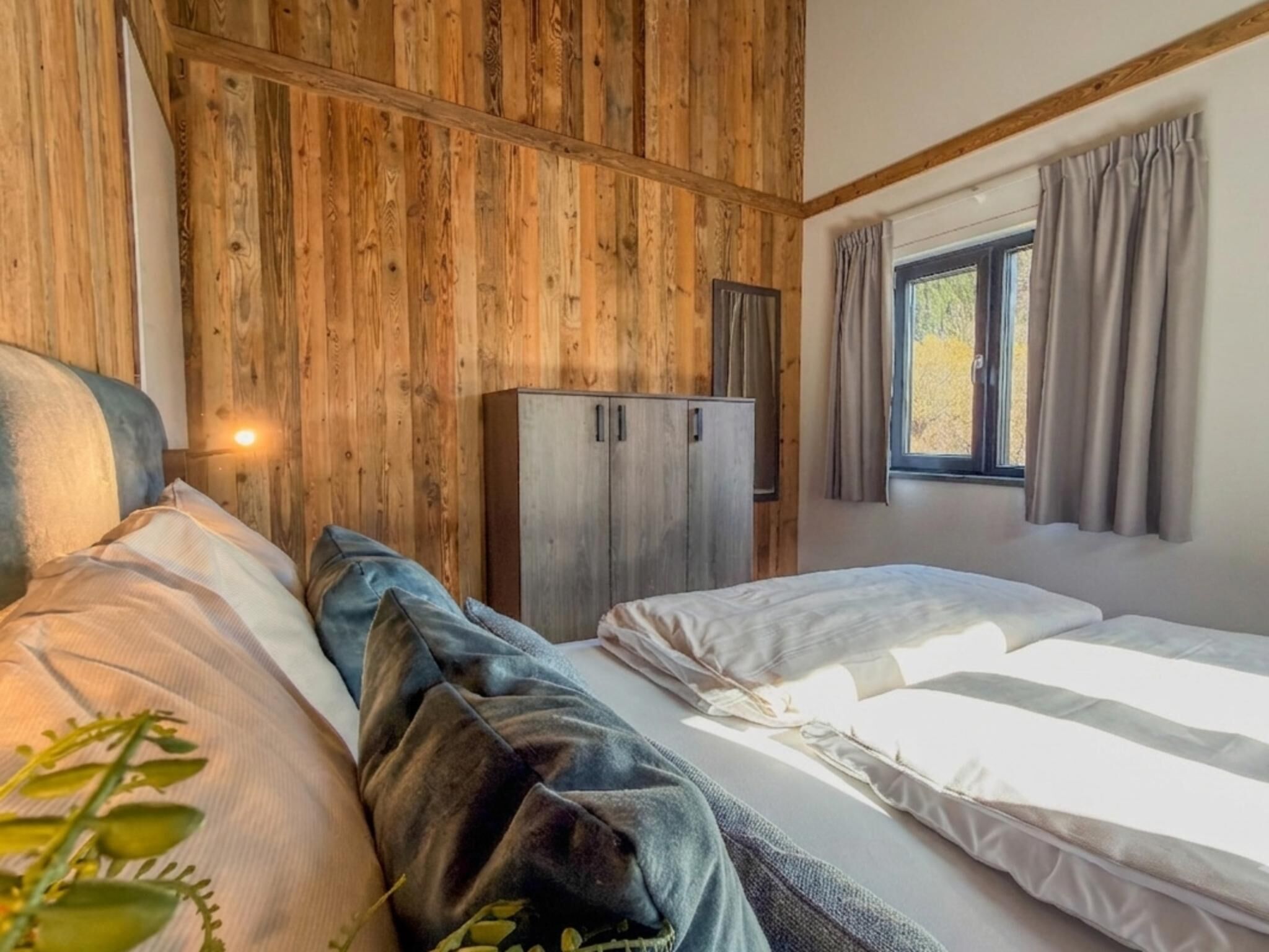 A bedroom with a wooden headboard, a wardrobe, a window with curtains, and a bed with pillows.