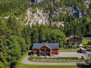 A two-story wooden cabin with a sloped roof, surrounded by dense forest and a rocky mountain backdrop.