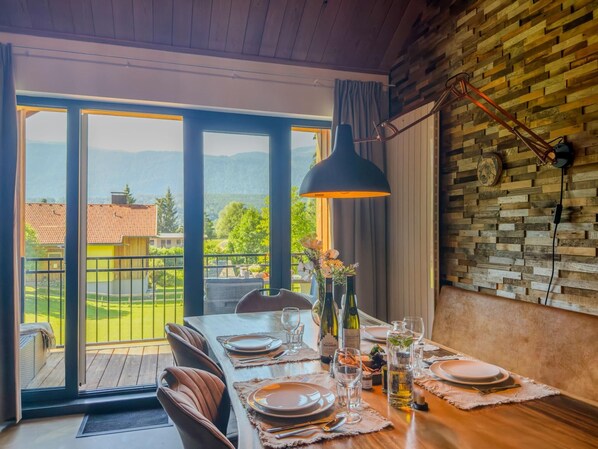 A dining area with a table set for two, a view of the outdoors, and a wooden wall with a clock.