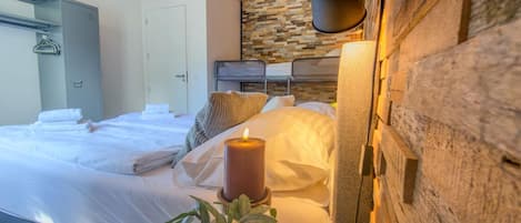 A bedroom with a wooden ceiling, a stone wall, a bed with white linens, a lit candle, and a potted plant.
