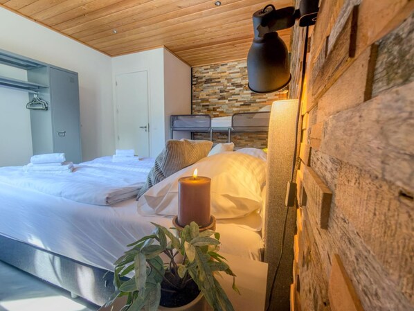 A bedroom with a wooden ceiling, a stone wall, a bed with white linens, a lit candle, and a potted plant.
