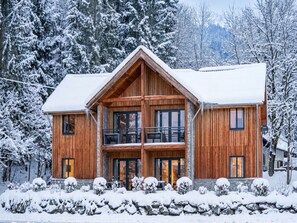 A two-story wooden cabin with a balcony on the upper floor, surrounded by snow-covered trees.