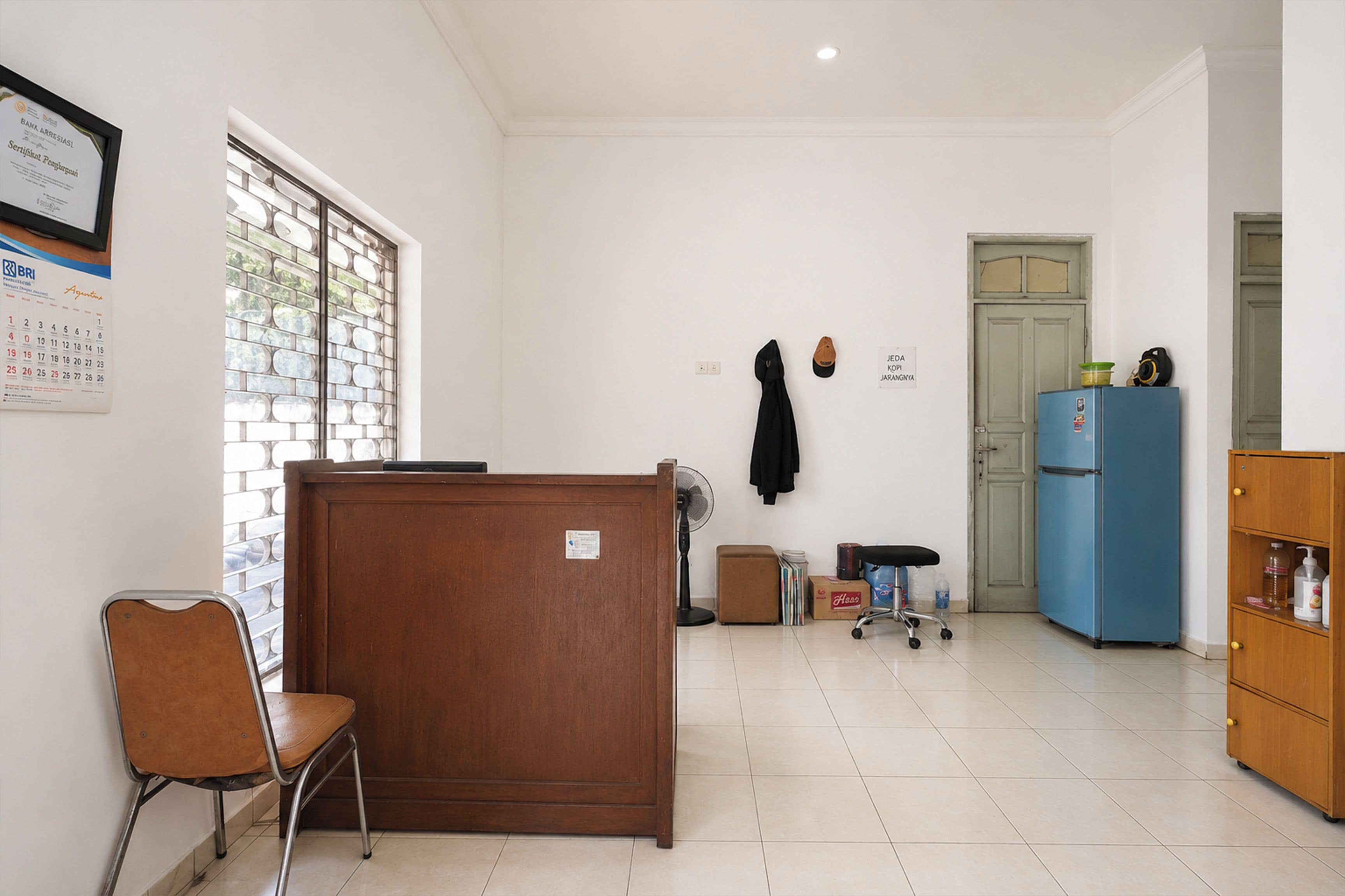 A room with a wooden desk, a chair, a blue refrigerator, and a door with a sign.