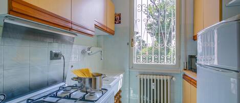 Una cocina compacta con estufa a gas, una olla de espagueti, una ventana con reja de metal y un refrigerador blanco.