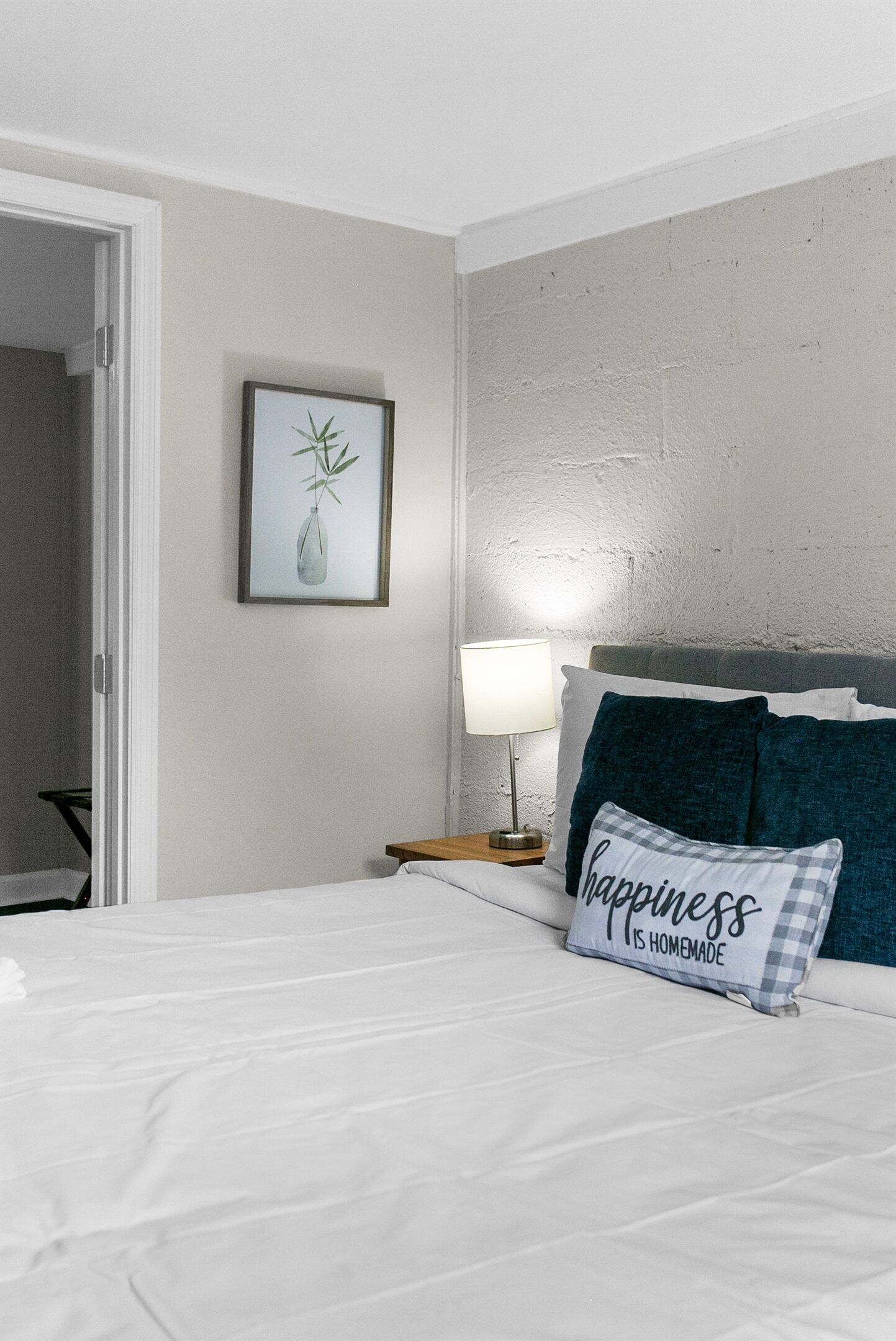 A neatly made bed with white bedding, a pillow with 'happiness is homemade' text, a bedside lamp, and a framed picture of a plant on the wall.