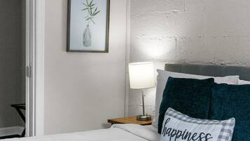 A neatly made bed with white bedding, a pillow with 'happiness is homemade' text, a bedside lamp, and a framed picture of a plant on the wall.