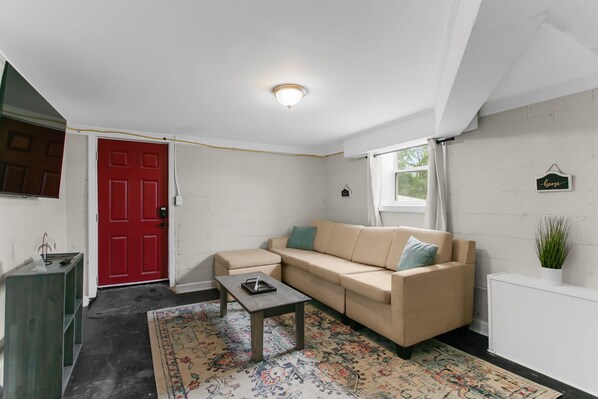 A living room with a beige sofa, a small table, a red door, and a white wall with a 'home' sign.