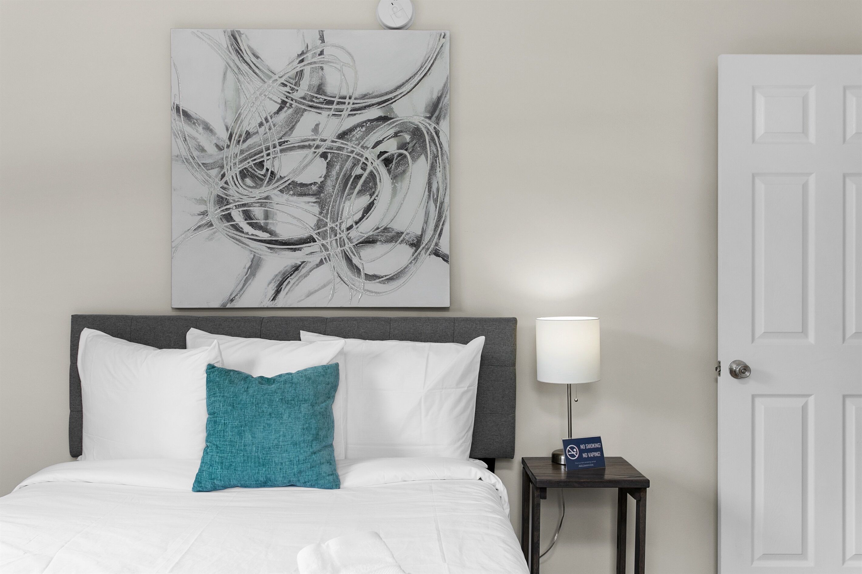 Une chambre d’hôtel avec un lit, une table de chevet avec une lampe, un panneau « Il est interdit de fumer » et un tableau abstrait accroché au mur.