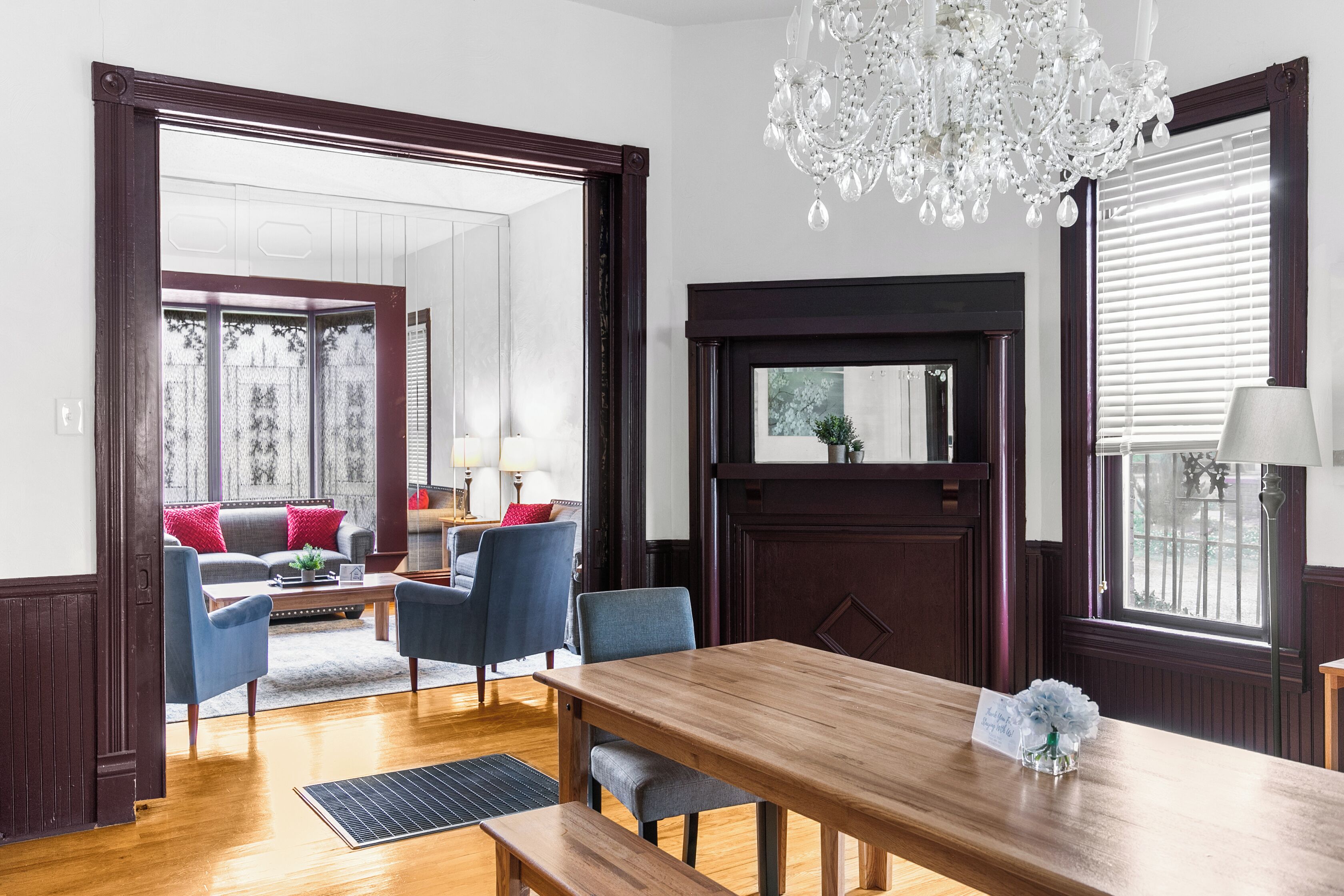 A spacious living room with a wooden dining table, blue chairs, a chandelier, and a large window with blinds.