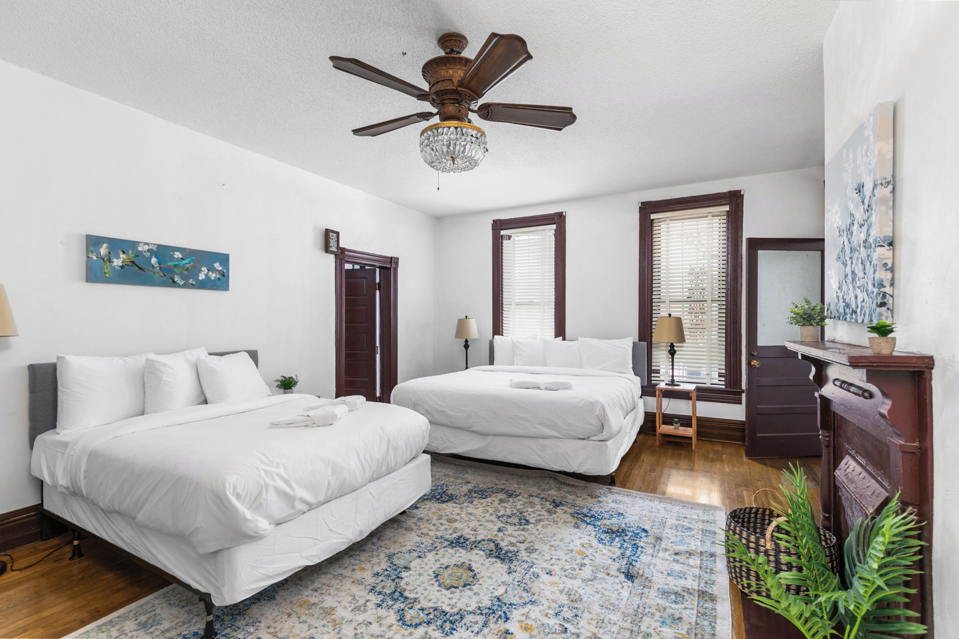 A hotel room with two beds, a ceiling fan, a chandelier, a painting, and a window with blinds.