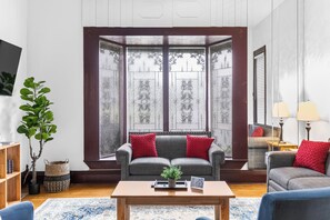 Ein Wohnzimmer mit Erkerfenster, einem grauen Sofa mit roten Kissen, einem Holztisch und einer Topfpflanze.
