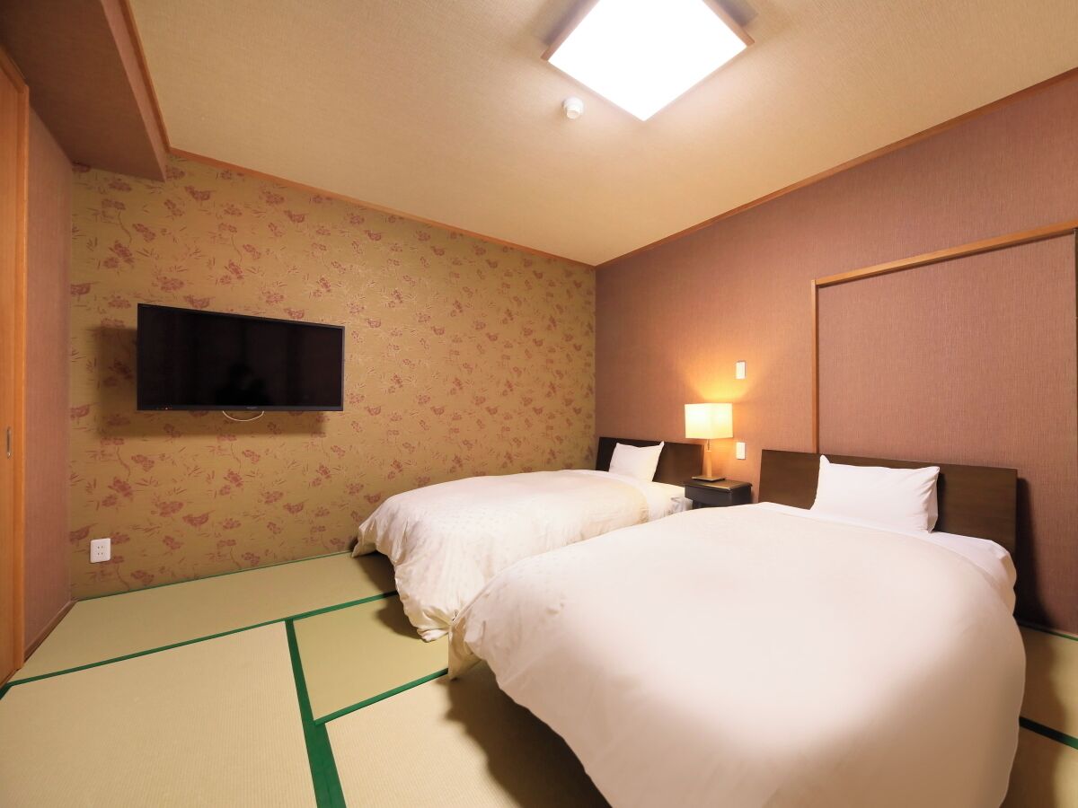 A hotel room with two beds, a flat-screen TV mounted on the wall, and a bedside table with a lamp.