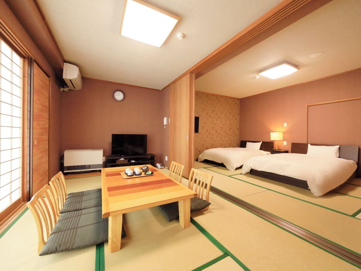 Habitación de hotel con suelo de tatami, una mesa de comedor con sillas, un televisor, dos camas y una mesita con juego de té.