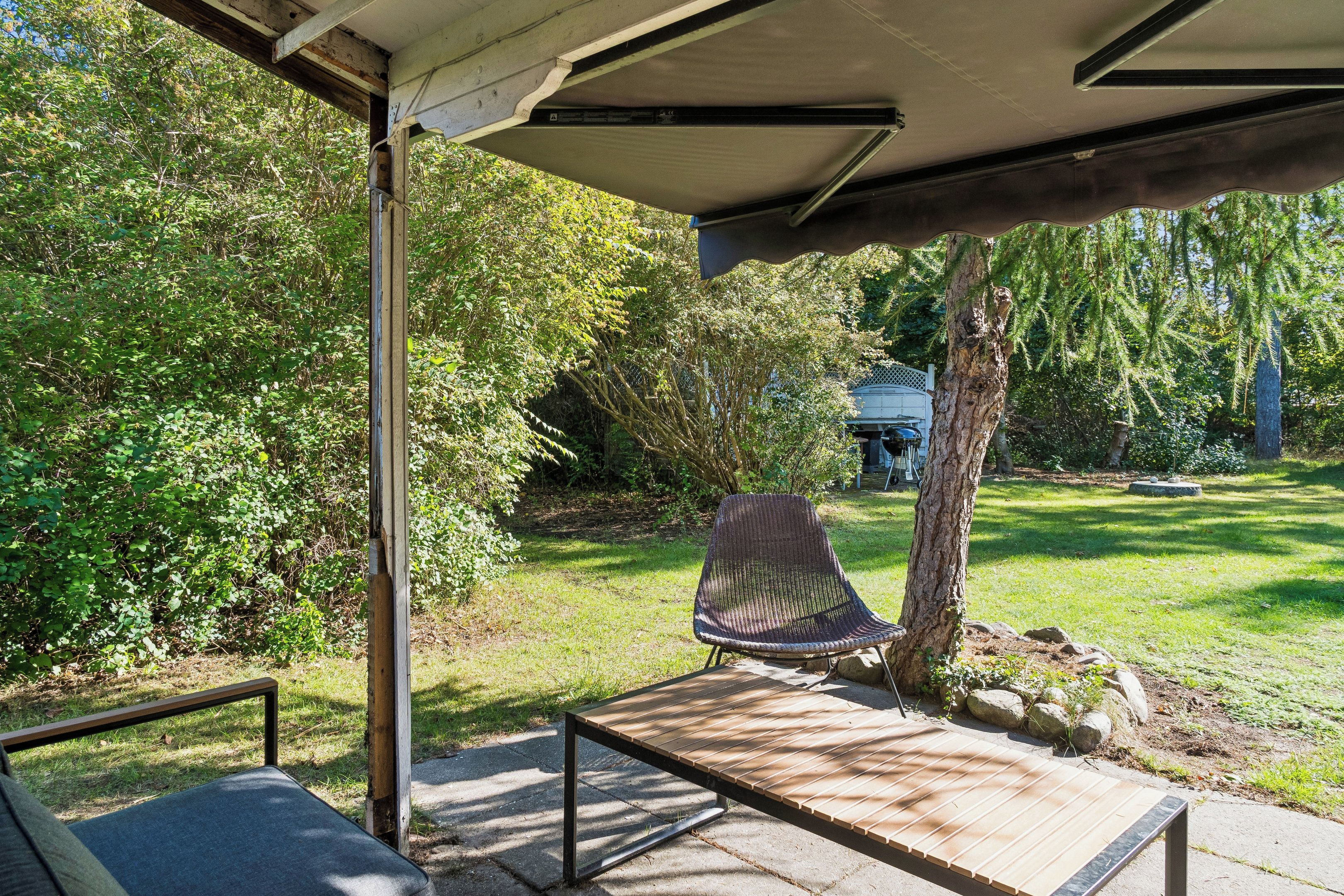 Un patio al aire libre con un banco de madera, una zona de estar cubierta y vistas a un jardín con árboles y una casa al fondo.