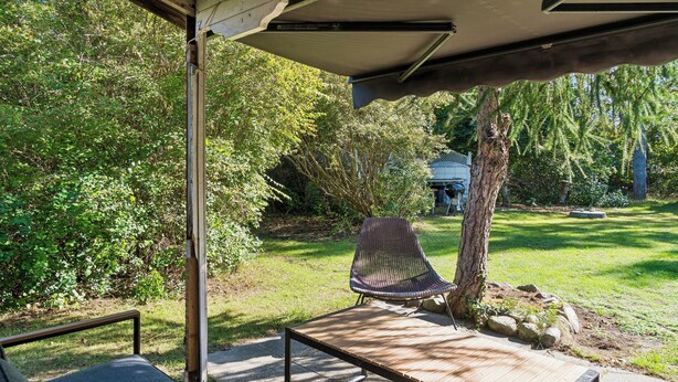 An outdoor patio area with a wooden bench, a covered seating area, and a view of a garden with trees and a house in the background. - Charming holiday home in beautiful Asserbo (Liseleje)
